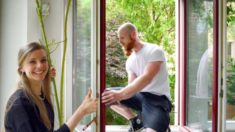 Eine Frau zeigt lächelnd den Daumen hoch, während ein Mann auf der Fensterbank ein Fenster öffnet oder einbaut. Im Hintergrund sind grüne Bäume sichtbar.