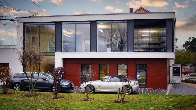 Ein modernes, zweistöckiges Gebäude mit großen Glasfronten und Holzverkleidung im Erdgeschoss. Vor dem Haus parken ein silbernes Cabrio und ein dunkleres Auto, umgeben von einer gepflegten Wiese und kleinen Bäumen.