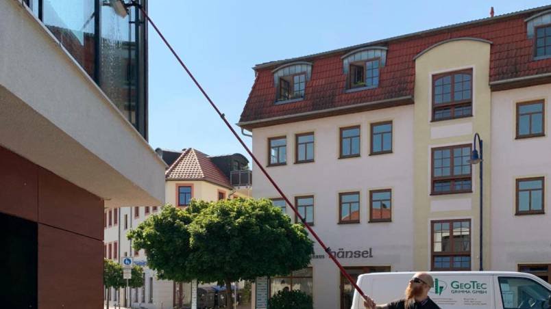 Martin Taxweiler reinigt mit einer langen Teleskopstange ein hohes Fenster an einem modernen Gebäude. Im Hintergrund sind klassische Häuser mit roten Dächern und ein grüner Baum zu sehen.