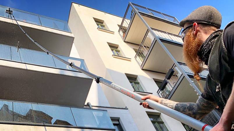 Ein Arm hält eine Teleskopstange, die zu einem Balkon eines modernen, mehrstöckigen Gebäudes mit Glasgeländern und weißen Wänden zeigt. Der Himmel ist blau.